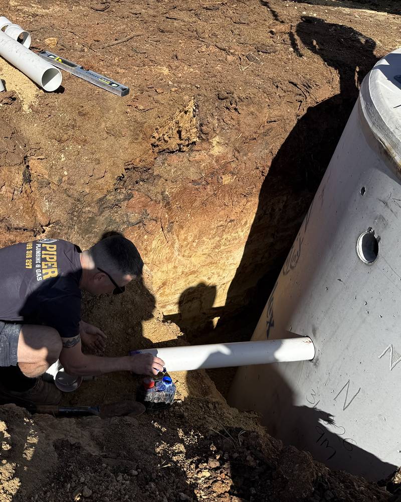 Craig kneeling in a dug-out hole, joining a white PVC pipe to a new concrete septic tank