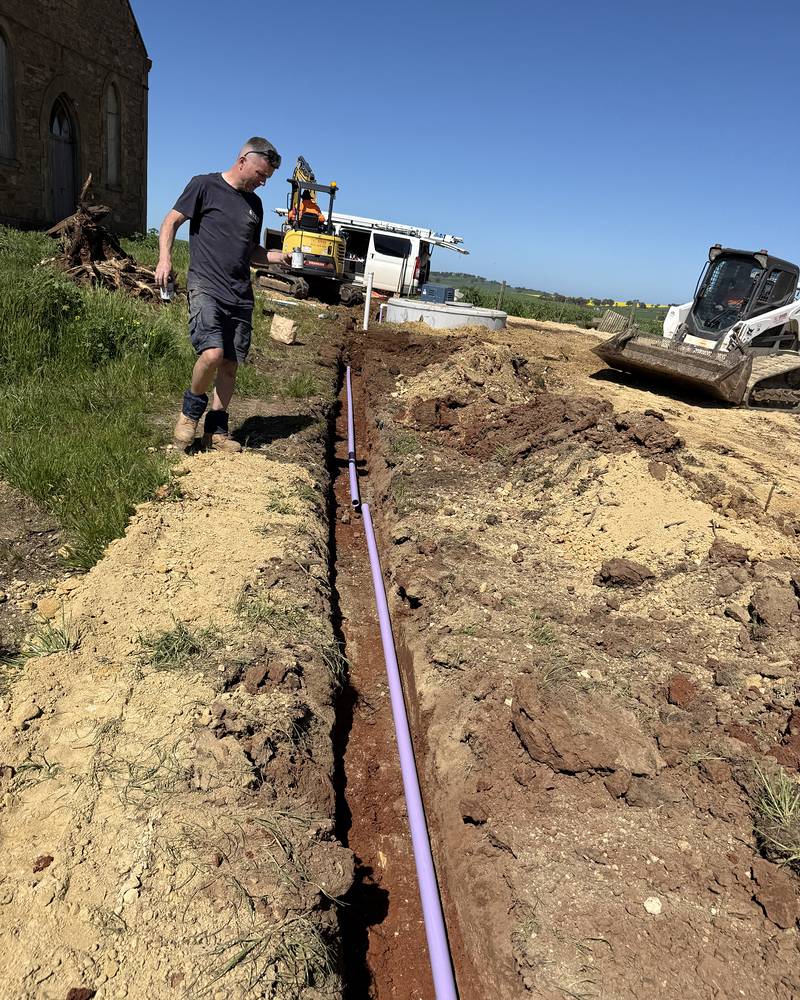 Craig walking beside a new wastewater line in a trench next to an old stone church