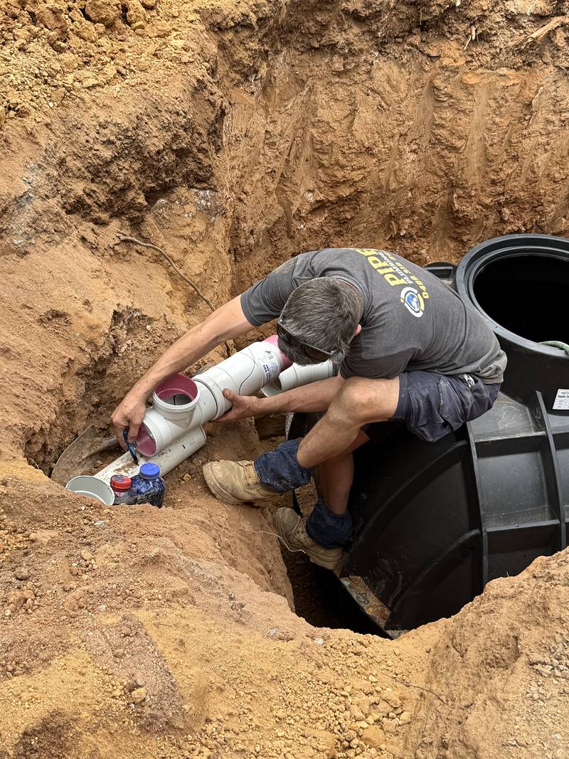 Craig kneeling on a new black septic tank in a red-soil hole, joining white PVC pipework to it