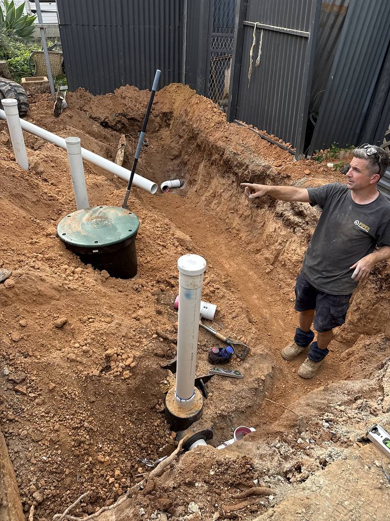 Open trench with white PVC risers and the green tank lid, Craig standing at the edge