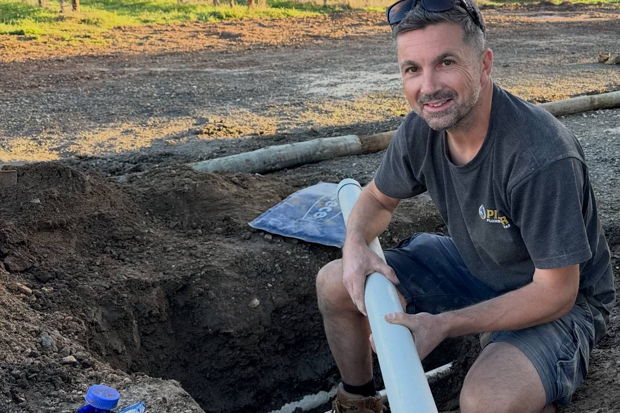 Craig kneeling in a trench beside a vineyard, holding a length of new PVC pipe