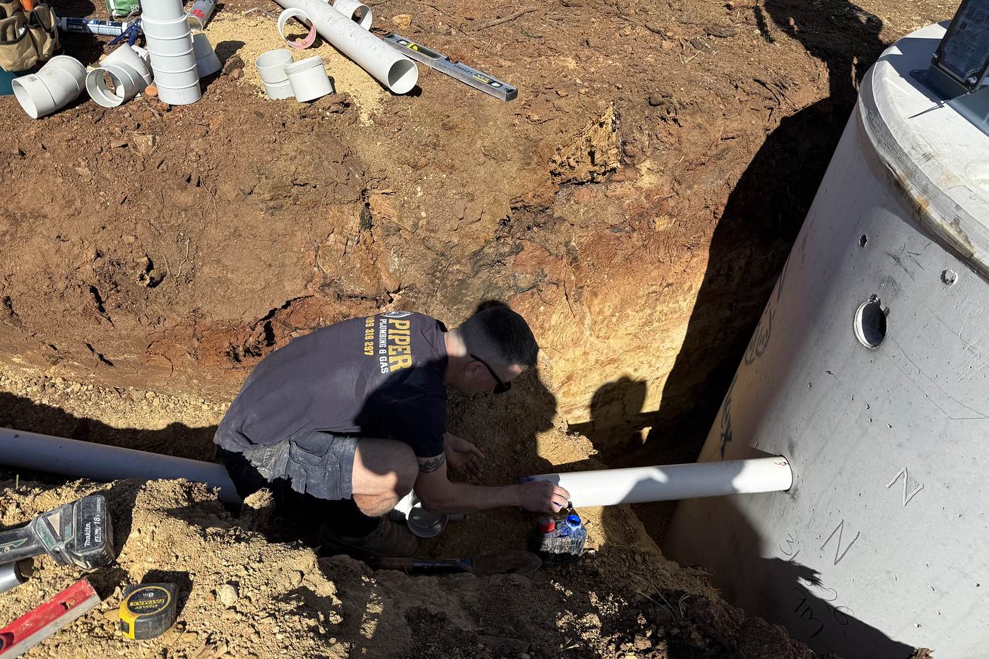 Craig kneeling in a dug-out hole, joining a white PVC pipe to the side of a new concrete septic tank