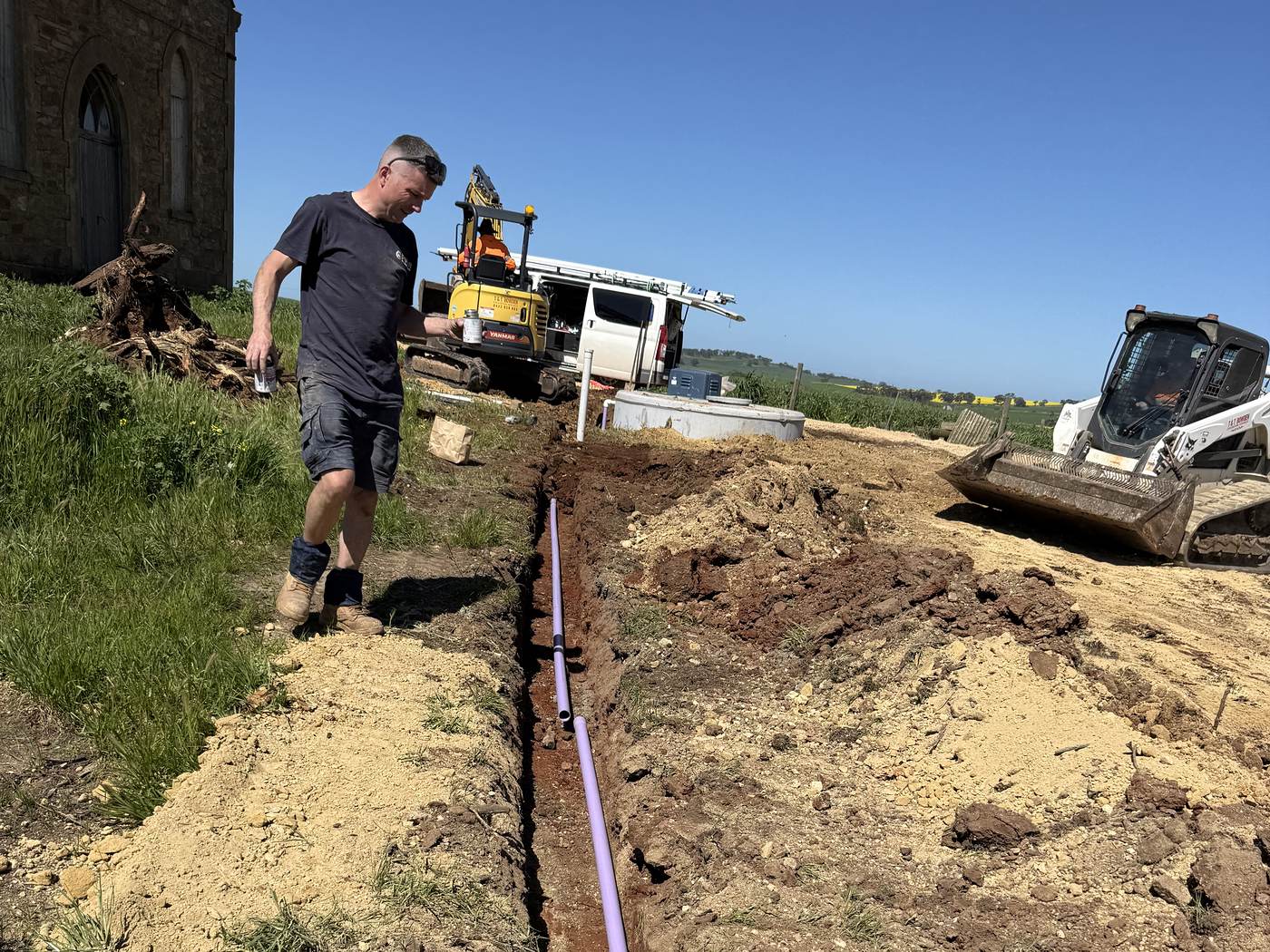 Craig walking beside a new wastewater line in a trench next to an old stone church, with an excavator and a bobcat behind