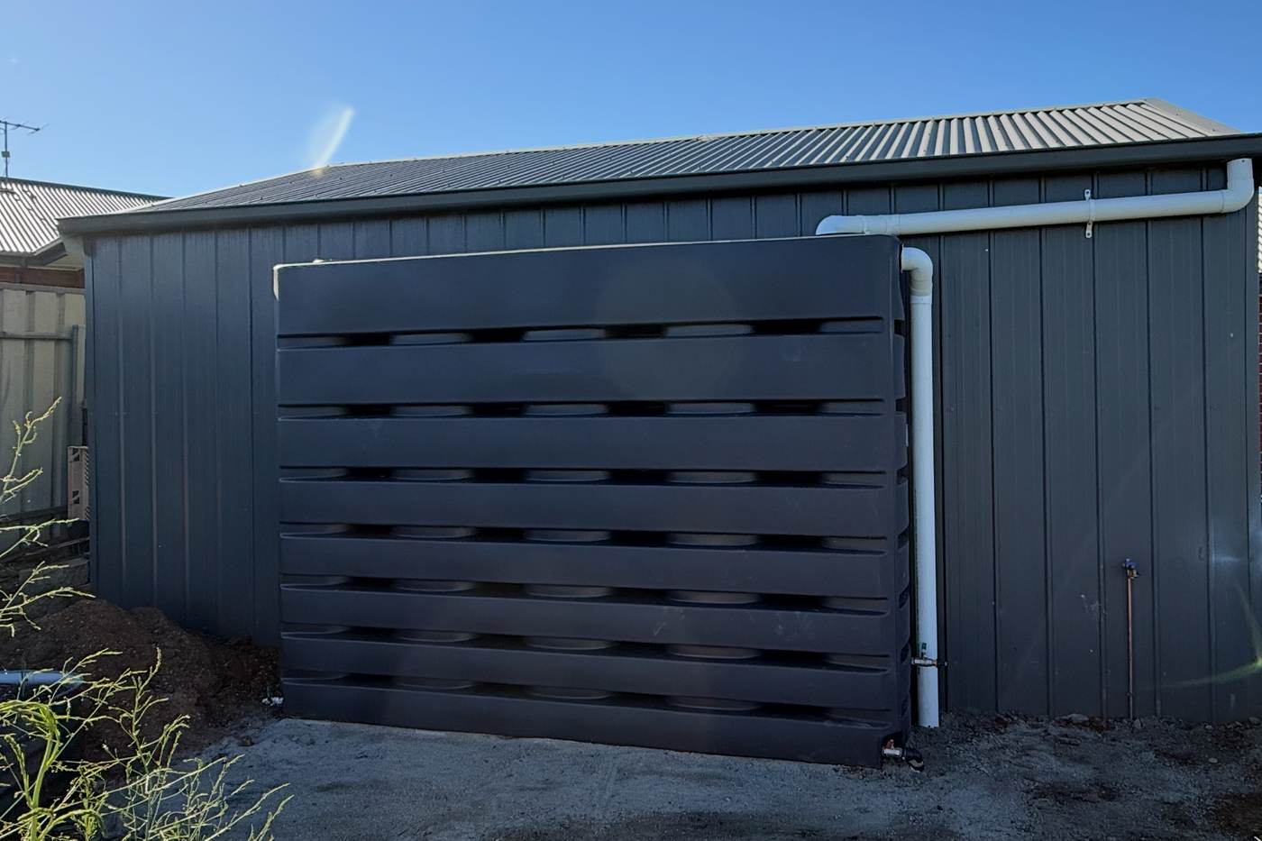 Dark slatted screen in front of a rainwater tank beside a shed, with new white downpipes