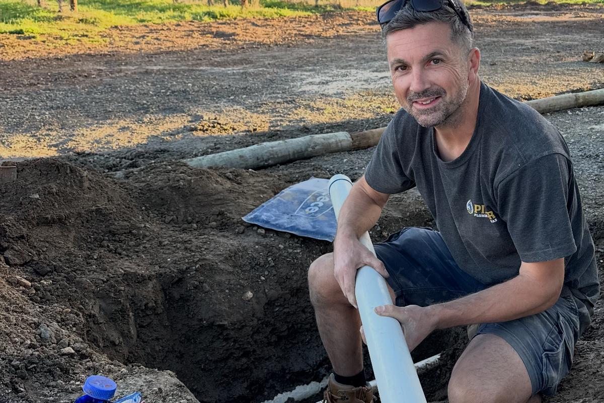 Craig holding a length of new PVC pipe in a trench beside a vineyard